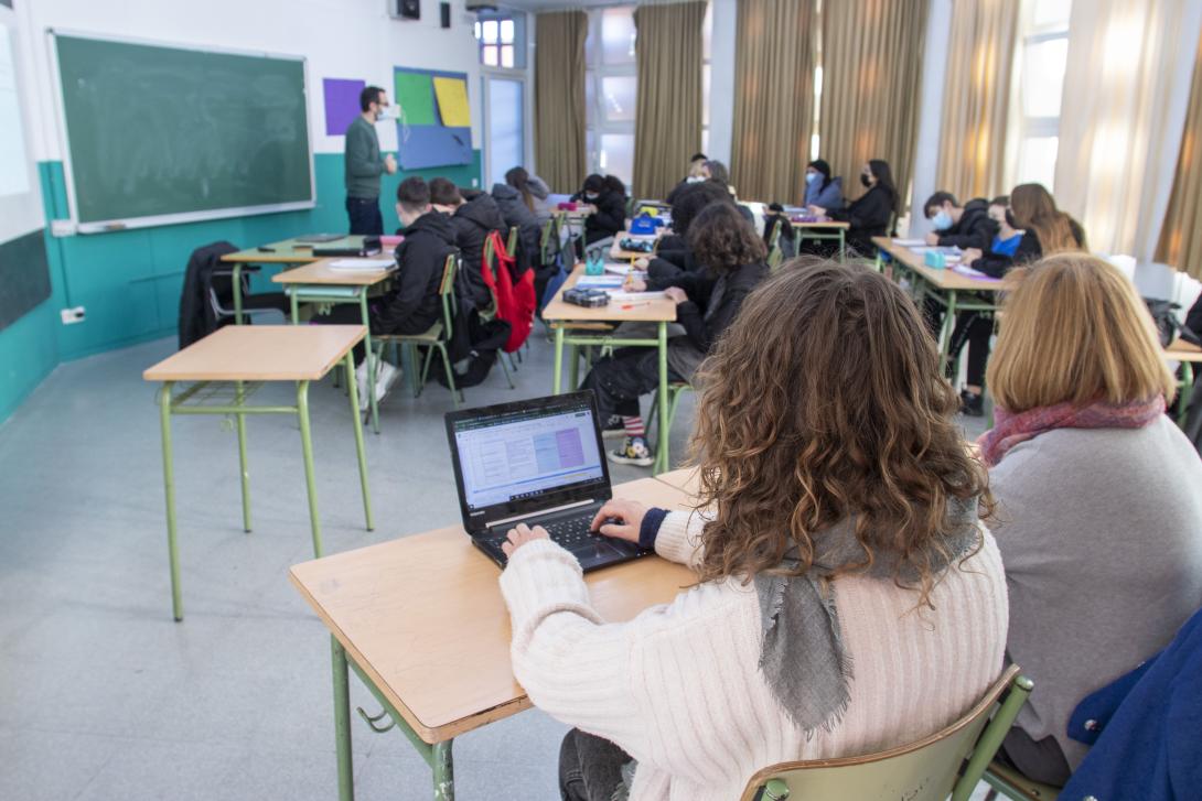 In the foreground, a woman uses a laptop computer while in the background a male teacher stands in front of a class of teenagers