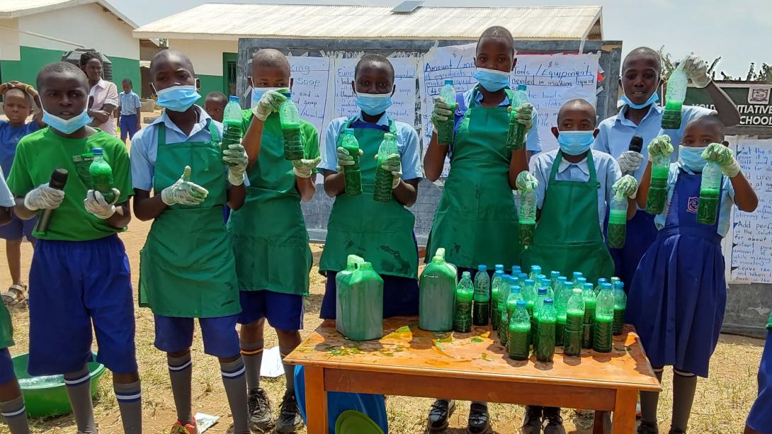 A group of Ugandan children in school uniforms and aprons hold up bottles filled with liquid soap