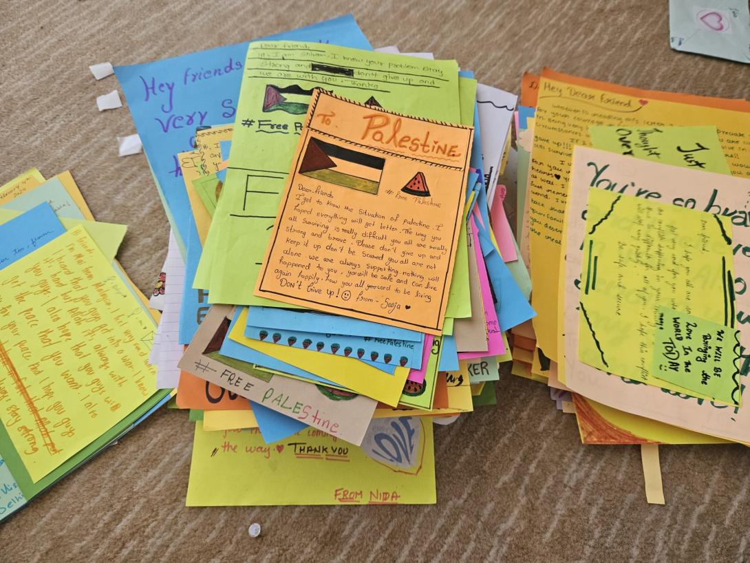 A hand-written letter on orange paper with the word Palestine at the top and a drawing of a Palestinian flag sits atop a pile similar letters on colorful paper