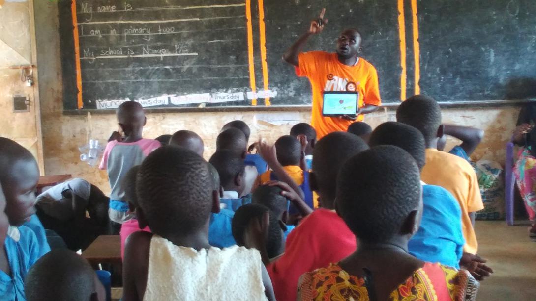 A teacher in an orange Teach For Uganda shirt gesticulates in front of a chalkboard in a classroom full of Ugandan students