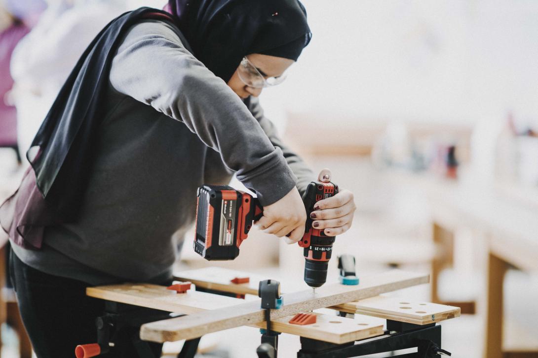 A teenage girl wearing a black head scarf, a grey sweatshirt and goggles uses a power drill on a plank of wood