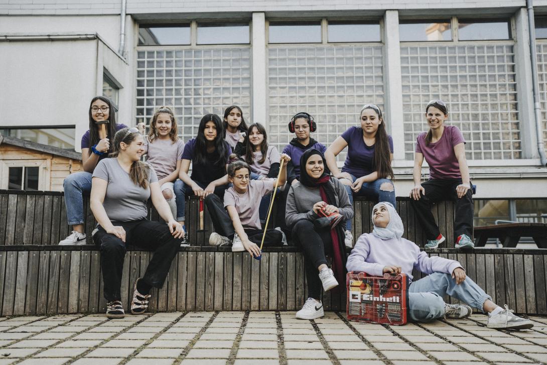 A young female teacher with a sandy blond ponytail smiles at a group of young teen girls posing while holding tools like hammars and drills