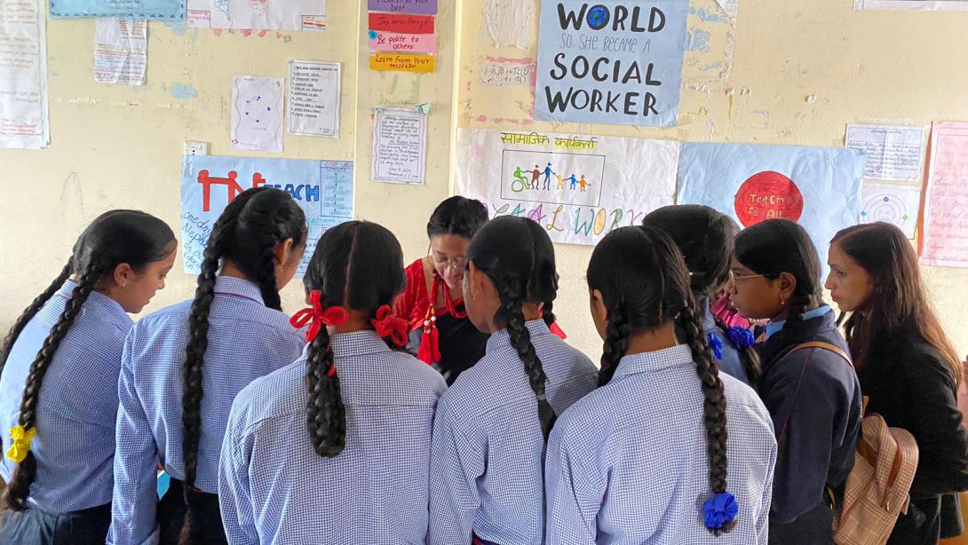 Girls in school unifrom huddle around a woman who is speaking, on the wall behind her are posters indicating she is a social worker