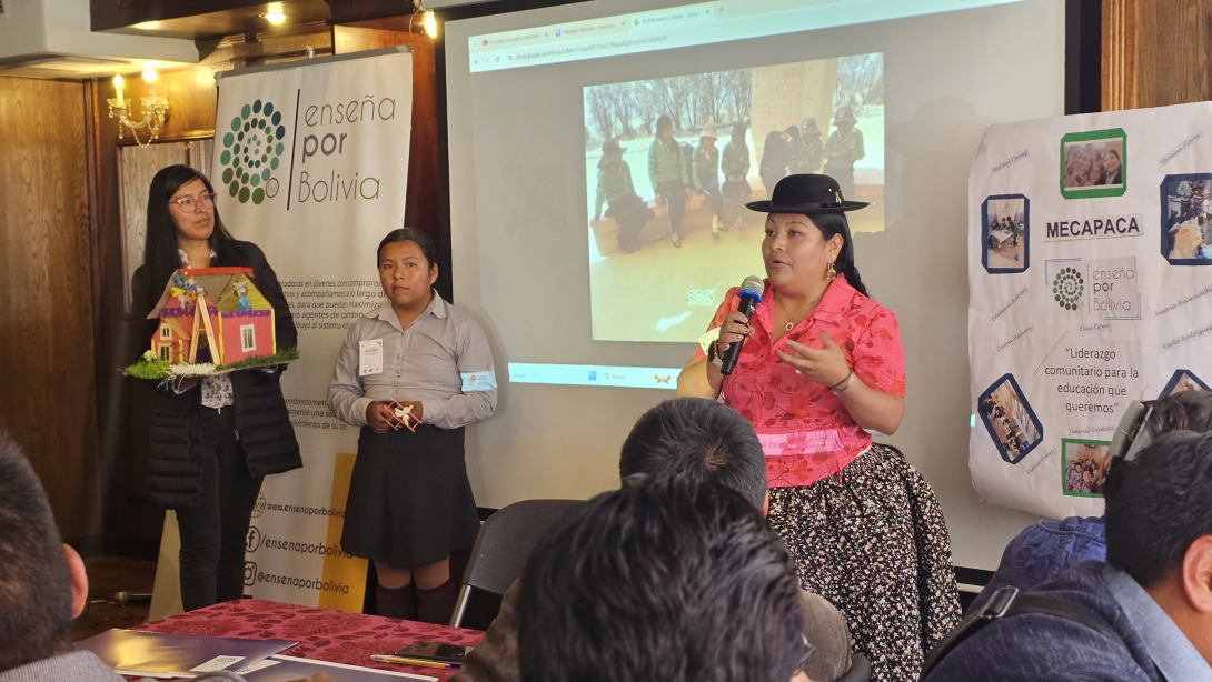 A Bolivian woman in traditional dress wearing a black rimmed hat does a presentation in front of a screen and a poster than says Mecapaca and Ensena por Bolivia