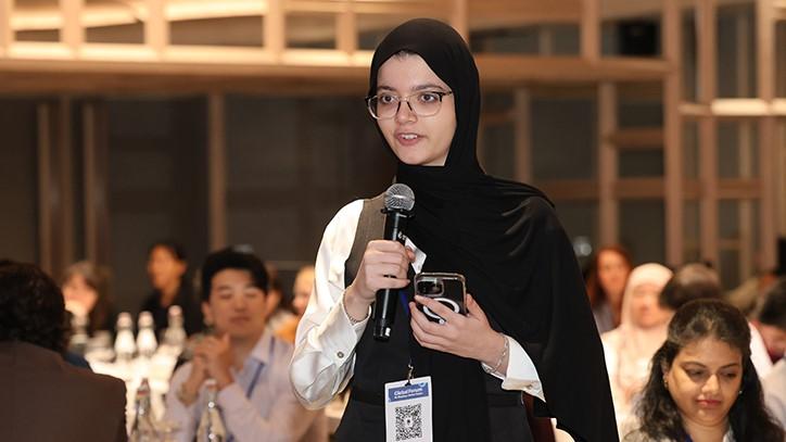 A young woman wearing a black head scarf and glasses speaks into a microphone 