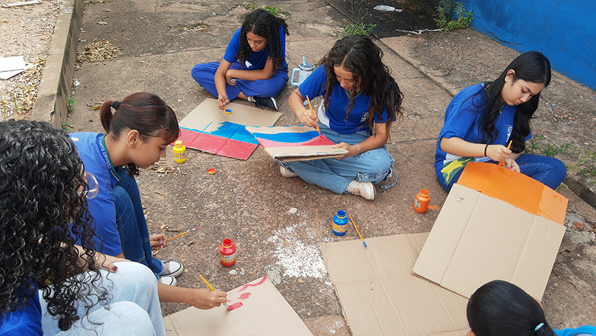 Five students in blue uniforms sit on the ground outdoors, painting colorful designs onto cardboard sheets using brushes and jars of bright paint