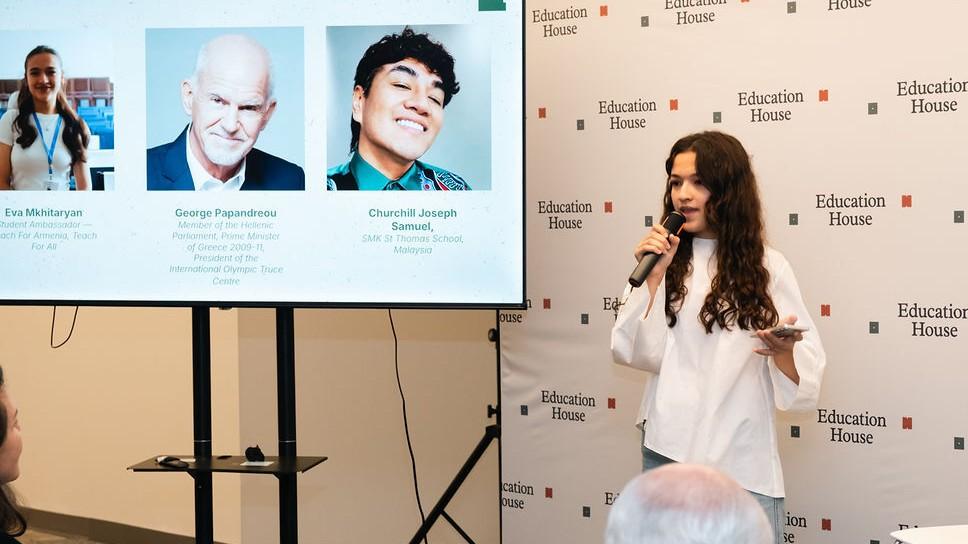 A young woman with light skin and long brown hair holding a microphone addresses an audience next to a screen displaying slide details and speaker headshots for "Education House"